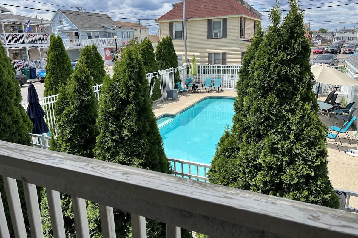 A swimming pool surrounded by tall trees, a white fence, and a building with a red roof.