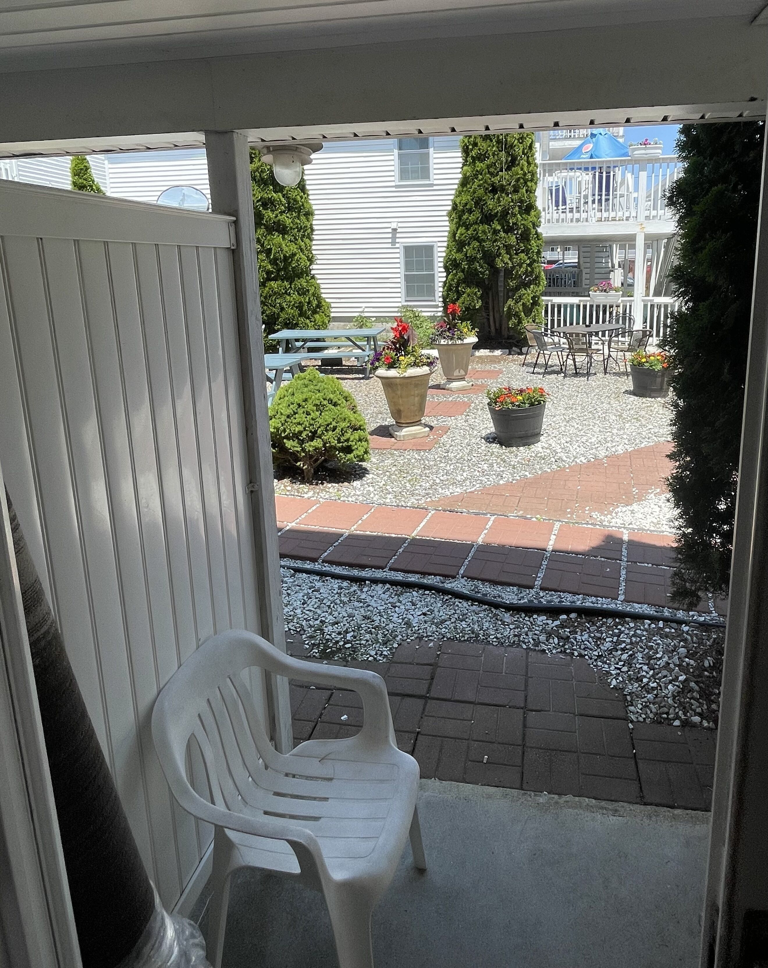 Een overdekt terras met een witte plastic stoel, planten in potten en een grindpad dat naar een gebouw met een balkon leidt.