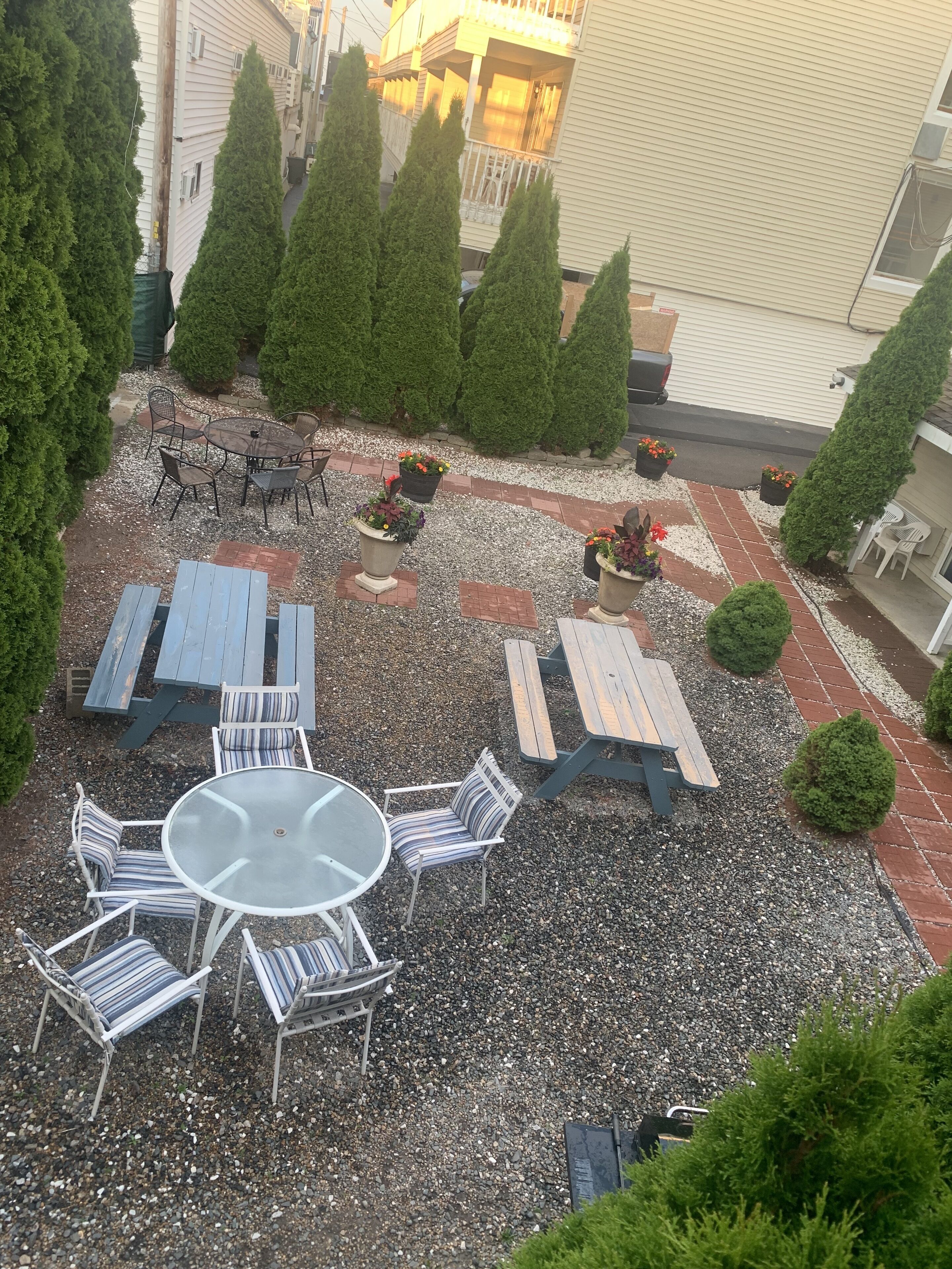 Un patio con mobiliario exterior, incluyendo mesas redondas y bancos, rodeado de plantas en macetas y árboles.
