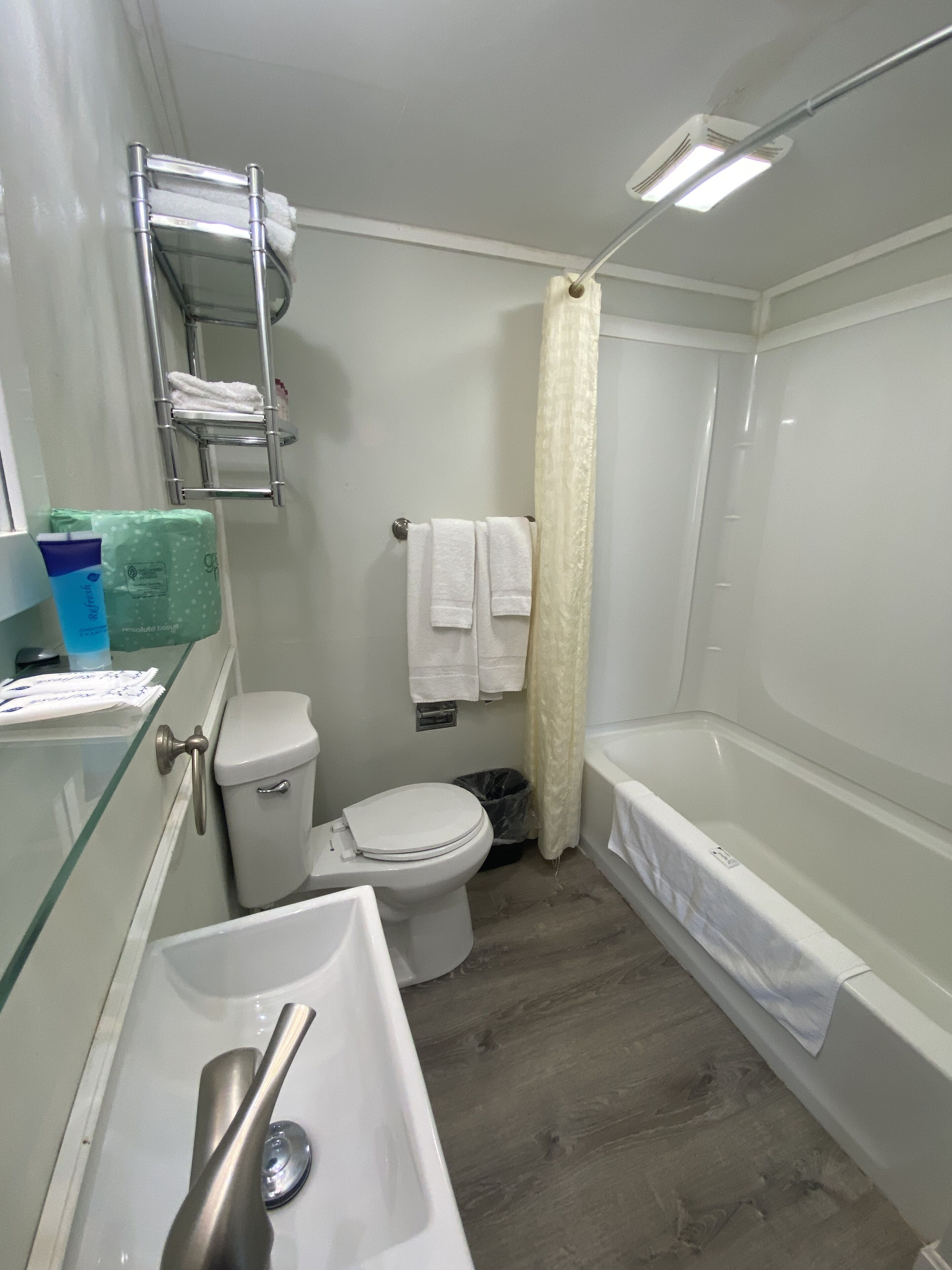 Baño con inodoro, lavamanos y bañera. Se observa un perchero con toallas y un cubo de basura.