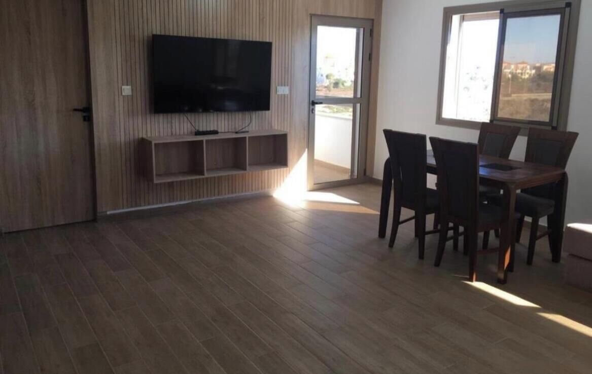 Ein Zimmer mit einem an einer Holzwand befestigten Flachbildfernseher, einem Esstisch mit Stühlen und einem großen Fenster mit Blick nach außen.