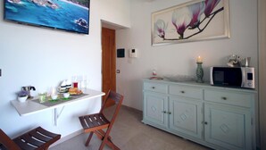 Ein Zimmer mit Fernseher, der eine Strandansicht zeigt, einem Esstisch mit Frühstücksgebäck, einem Sessel, einem Schrank mit Mikrowelle und einem Blumenbild.