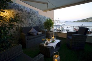 Een balkon met een bank, stoelen, een tafel met drankjes, een potplant en uitzicht op zee en boten.