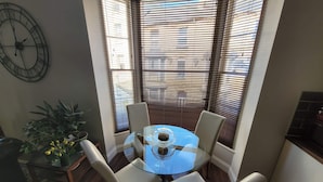 A corner dining area with a round glass table, four chairs, a potted plant, and a wall clock.