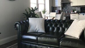 A living room with a black leather sofa, a large wall clock, and a kitchen in the background.