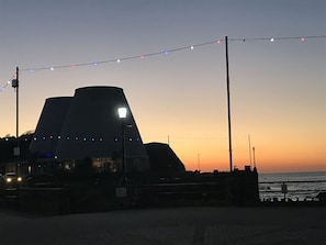A coastal building silhouette against a sunset sky, with string lights overhead and a paved walkway leading to the water.