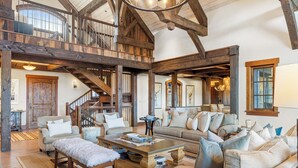 Ein geräumiges Wohnzimmer mit hoher Decke, freiliegenden Holzbalken, einer Treppe und einer gemütlichen Sitzecke mit mehreren Sofas und einem Couchtisch.