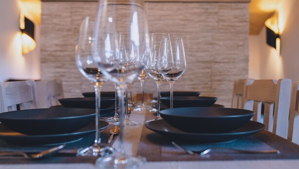 Une table à manger dressée avec de nombreux verres à vin et des assiettes, sur fond d’un mur en bois et de luminaires fixés au mur.