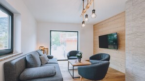 Un salon moderne avec un canapé gris, un fauteuil foncé, une table basse en bois et un téléviseur fixé à un mur en bois.