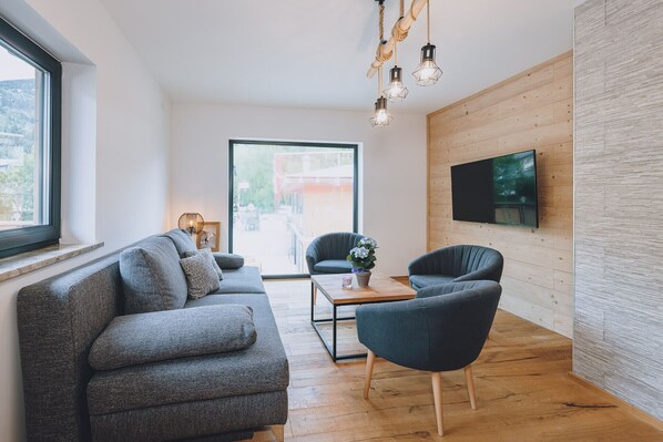 A modern living room with a grey sofa, a dark armchair, a wooden coffee table, and a TV mounted on a wooden wall. - Premium Apartments Areit - Balcony, sauna (Zell am See)