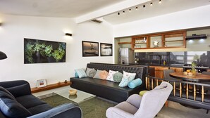 A modern living room with a black sofa, grey armchair, and a glass coffee table. A kitchen area with wooden cabinets and a dining table is visible in the background. - Villa Taleona (Cóbano)