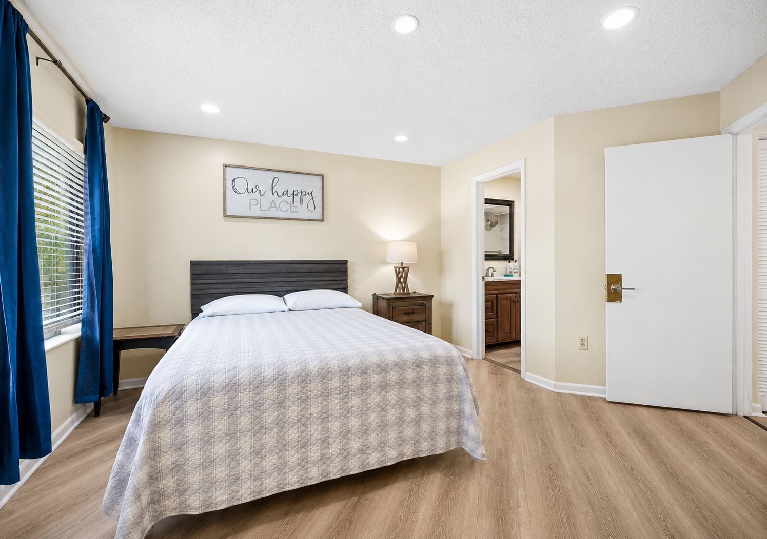 A hotel room with a bed, a nightstand, a bedside table with a lamp, a bathroom visible through an open door, and a sign on the wall.