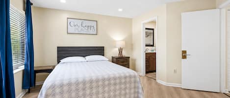 A hotel room with a bed, a nightstand, a bedside table with a lamp, a bathroom visible through an open door, and a sign on the wall.