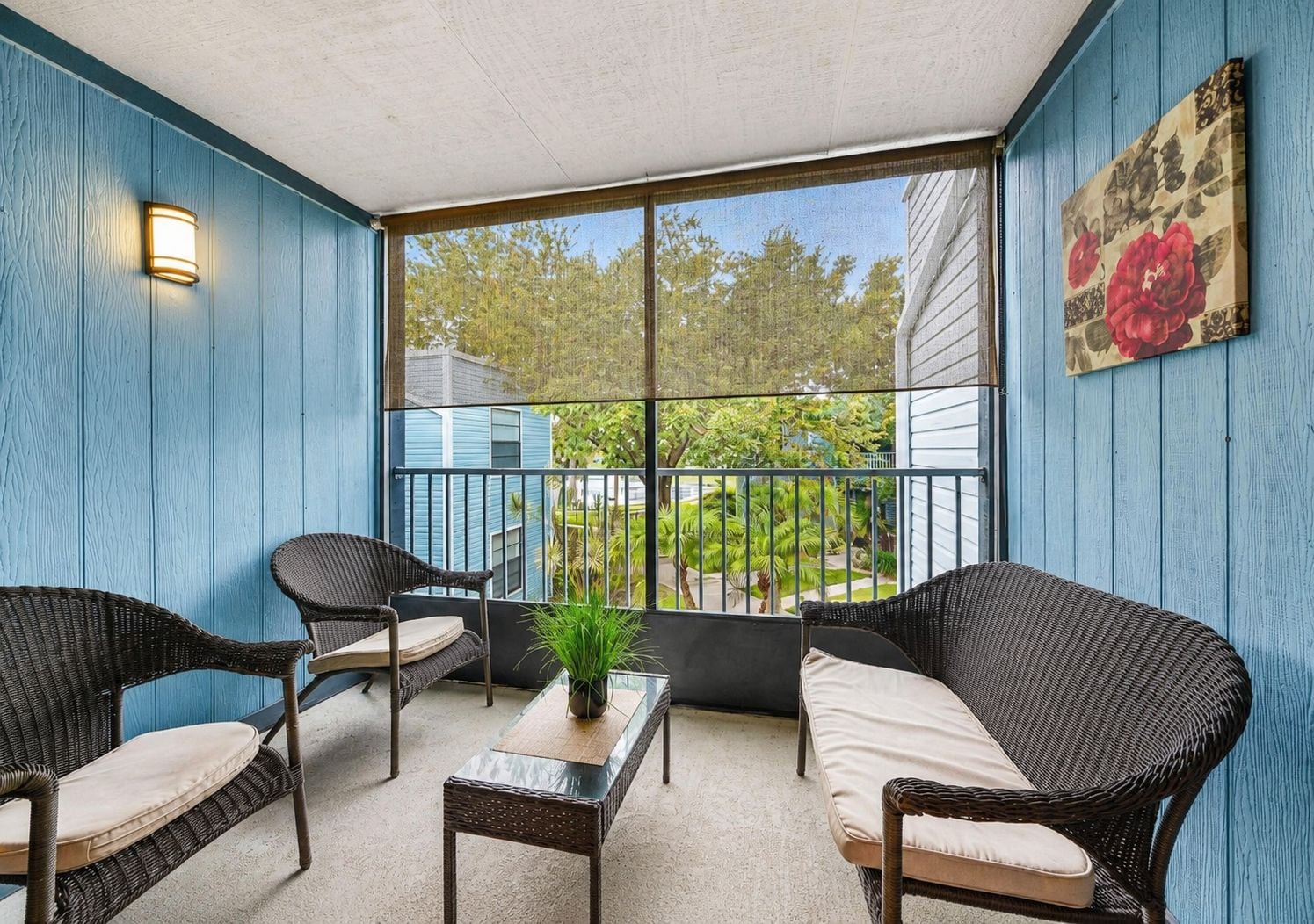 Una habitación con paredes azules, muebles de mimbre, una mesa de vidrio y vistas a un jardín y una casa.