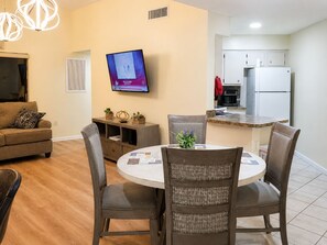 A modern living room with a round dining table, grey upholstered chairs, a flat-screen TV mounted on the wall, and a kitchen area with white cabinets and a refrigerator. - 3648 Orlando Vacational Apartment 2nd floor, Lake view (Orlando)