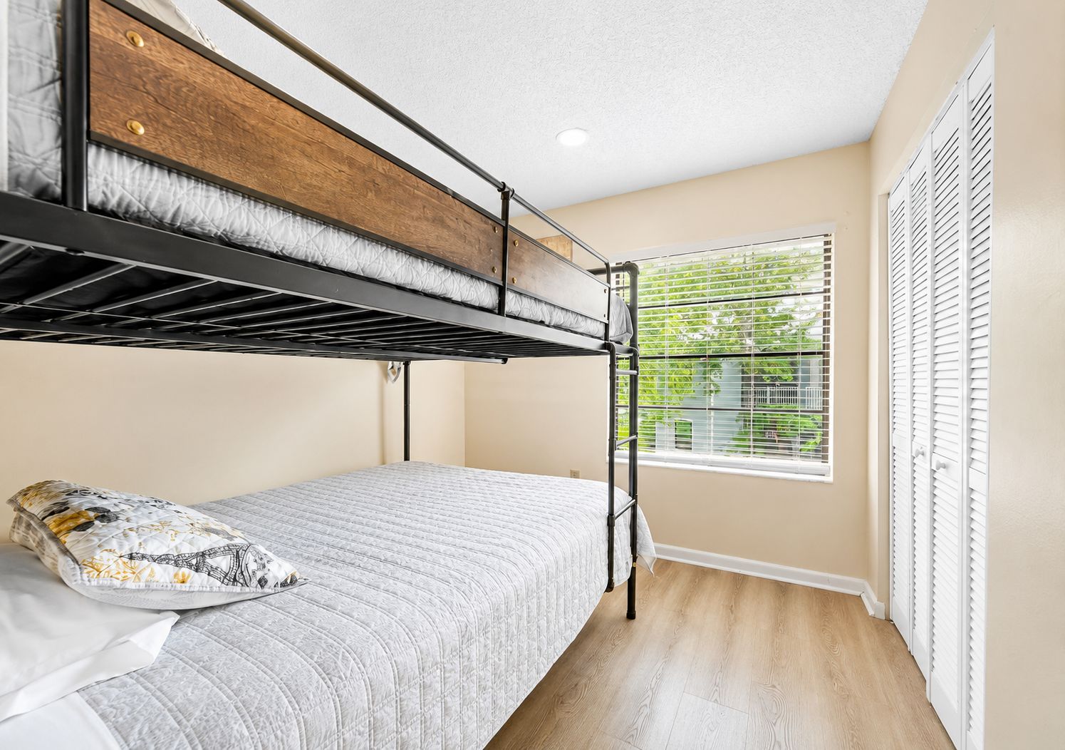 Una cama litera con cabecera de madera, una cama individual con almohada estampada, una ventana con persianas y una puerta con contraventanas.