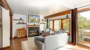 A spacious living room with a fireplace, a television mounted on the wall, and a large window with a view of the outdoors.