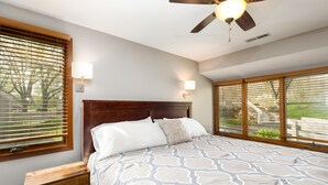 A bedroom with a bed, wooden headboard, two wall-mounted lamps, a ceiling fan, and large windows with blinds.