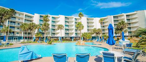 A hotel pool area with multiple lounge chairs and umbrellas, surrounded by palm trees and a large building with balconies.