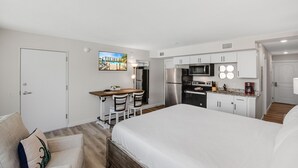 Hotellihuone, jossa on sänky, työpöytä tuoleineen, pieni keittiönurkkaus ja ovi.