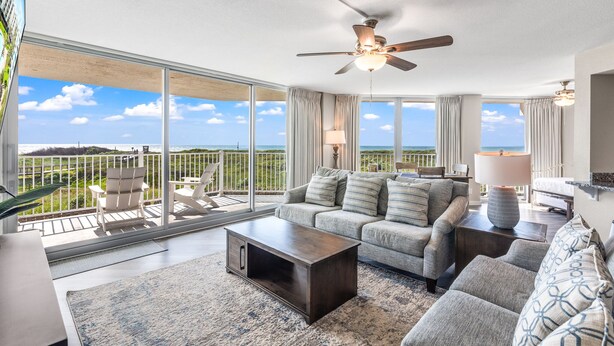 A spacious living room with a large window overlooking a scenic view, a ceiling fan, and a modern sofa set. - Sandcastle Condominium Unit 137 (Port Aransas)
