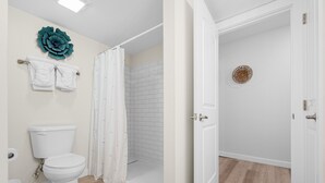 A bathroom with a toilet, shower, and a door leading to another room. - Sandcastle Condominium Unit 214 (Port Aransas)