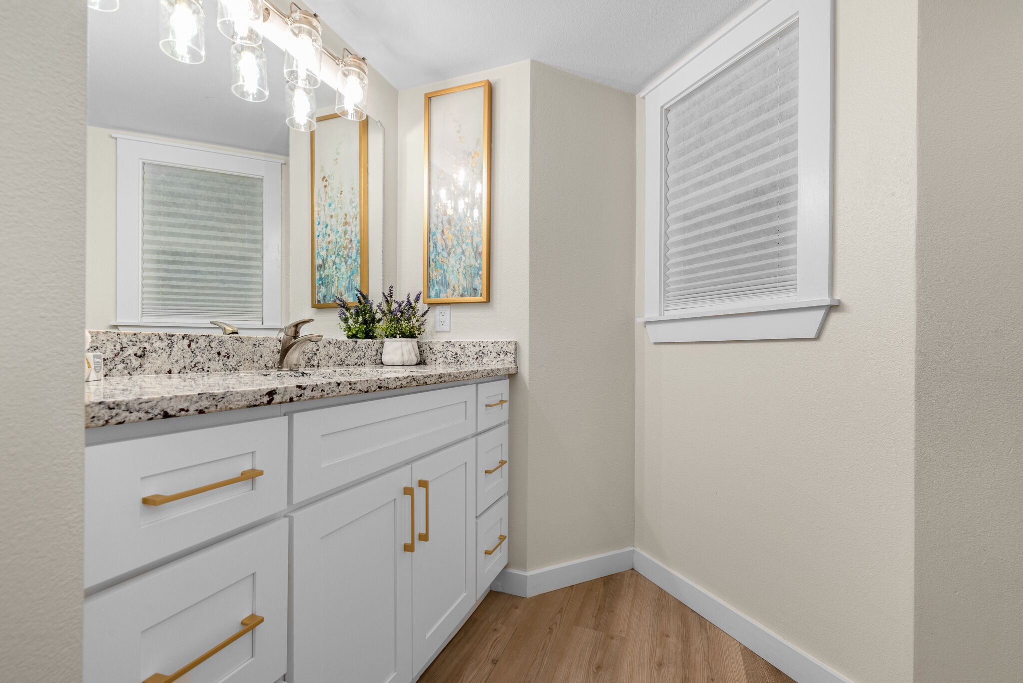 Une salle de bain avec des meubles blancs, un comptoir en granit, un lavabo, un miroir et une fenêtre avec des stores.