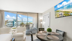 A modern hotel room with a balcony, a dining area with a round table and chairs, and a large window offering a view of the beach and buildings. - Sandcastle Condominium Unit 214 (Port Aransas)
