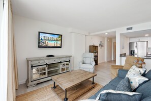 A living room with a wooden coffee table, a sofa with patterned cushions, a television mounted on the wall, and a view of a pool through the window. - Sandcastle Condominium Unit 220 (Port Aransas)