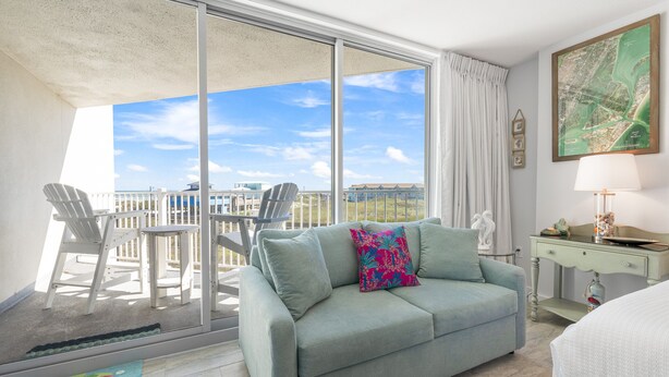 Een balkon met uitzicht op zee, een lichtblauwe bank met een kleurrijk kussen en een klein tafeltje met een lamp.