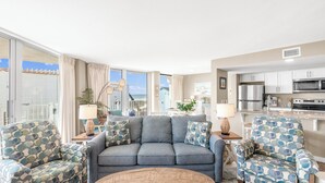 A modern living room with a grey sofa, patterned armchairs, a round coffee table, and a kitchen area with white cabinets and stainless steel appliances. - Sandcastle Condominium Unit 237 (Port Aransas)