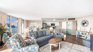 A modern living room with a grey sofa, patterned armchairs, a coffee table, and a kitchen in the background. - Sandcastle Condominium Unit 237 (Port Aransas)