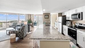 A modern kitchen with white cabinets, a granite countertop, and stainless steel appliances. There is a seating area with patterned chairs and a view of the ocean through large windows. - Sandcastle Condominium Unit 237 (Port Aransas)