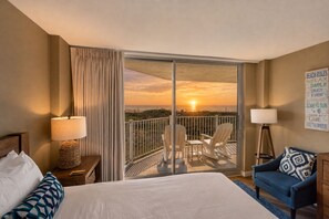 A hotel room with a bed, two chairs, a sofa, a lamp, and a view of the sunset over the ocean.