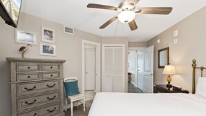 Une chambre à coucher comprenant un lit, une commode, une chaise, un ventilateur de plafond et une porte donnant sur une autre pièce.