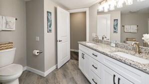 Une salle de bain avec des toilettes blanches, un lavabo doté d’un plan de travail en granit et un grand miroir au-dessus du lavabo.