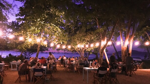 An outdoor dining area with tables and chairs under trees, overlooking a body of water. - Beachfront One Bedroom on Tamarindo Beach (Tamarindo)