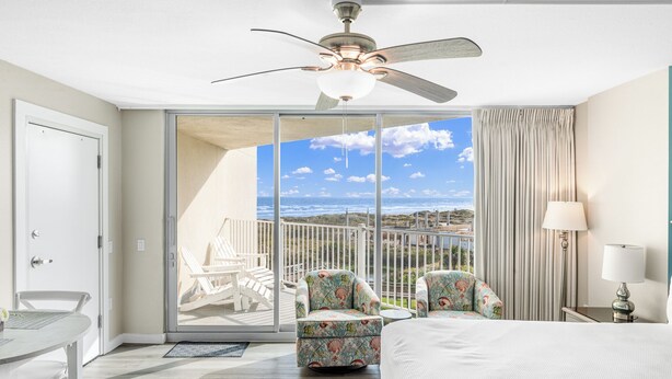 A hotel room with a bed, a ceiling fan, a view of the ocean, and a balcony with chairs. - Sandcastle Condominium Unit 311 (Port Aransas)