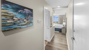 A hallway with a painting of a heron and water lilies, a white door, and a view into a bedroom with a bed and curtains. - Sandcastle Condominium Unit 311 (Port Aransas)
