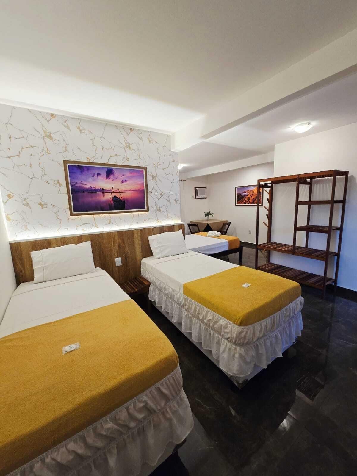 A hotel room with two single beds, a wooden headboard, a marble-patterned wall, a framed sunset picture, a small table with a chair, and a wooden shelf unit.