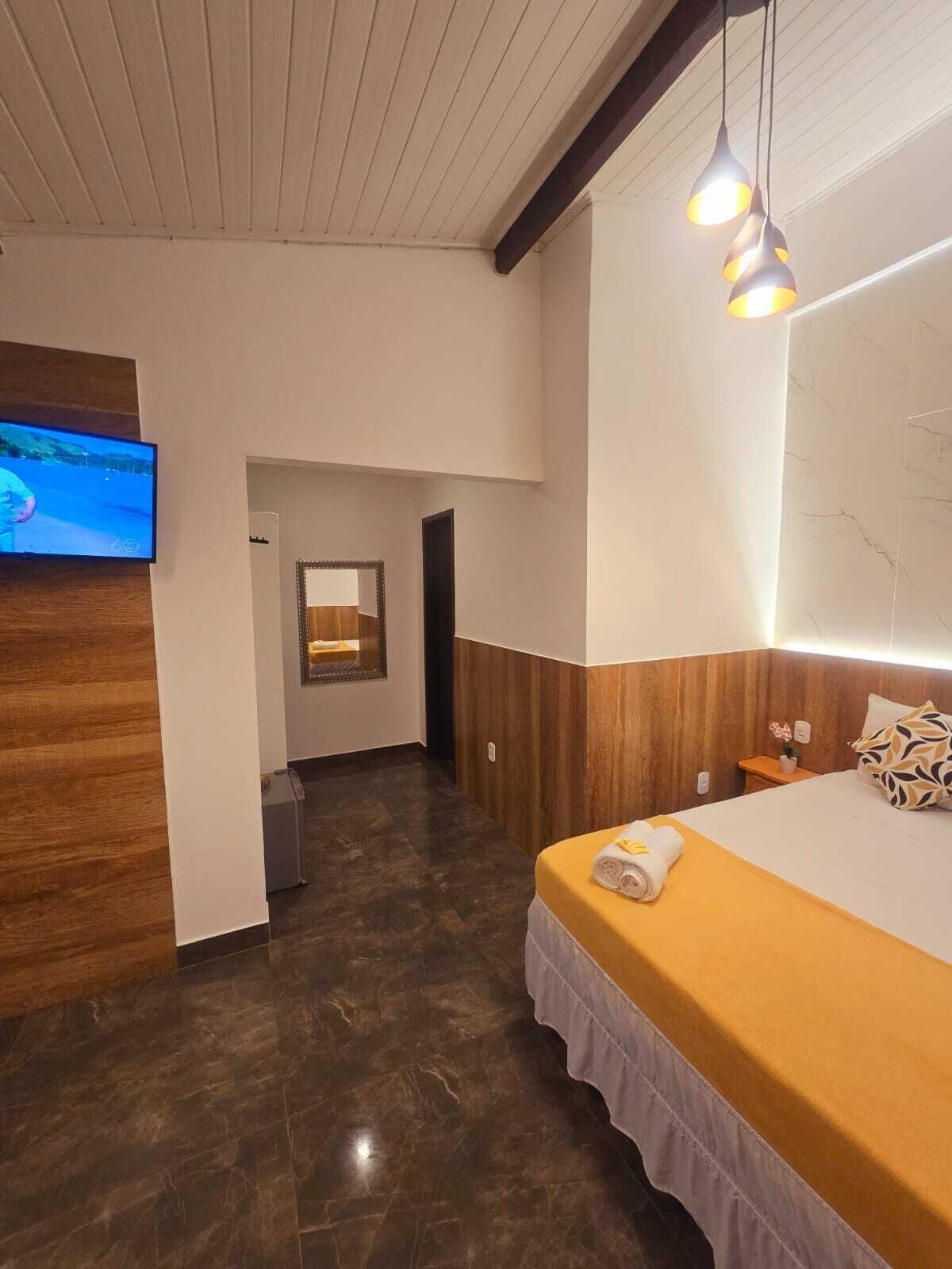 A hotel room with a bed, a TV mounted on the wall, a wooden accent wall, a mirror, and a hallway leading to another room.