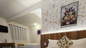 Habitación de hotel con cama, mesita de noche, televisión, un cuadro en la pared y ventana con cortinas.