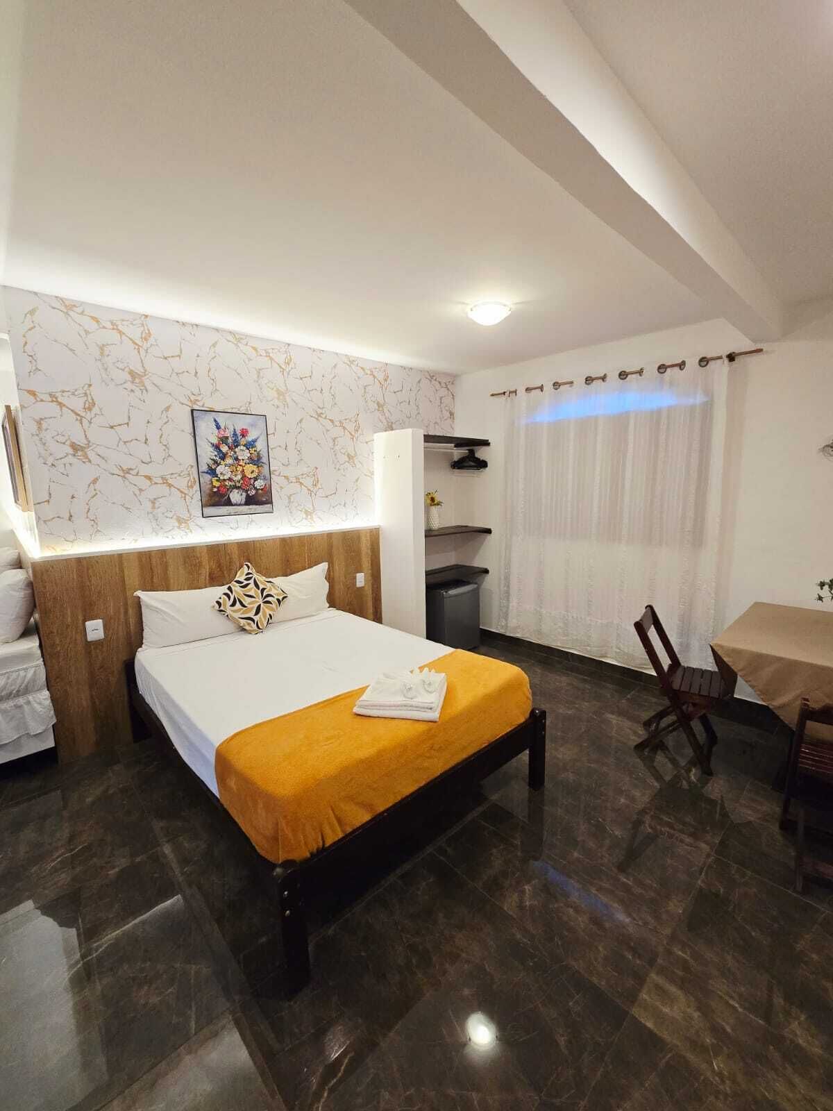 A hotel room with a bed, a small table, a chair, a picture on the wall, and a window with curtains.
