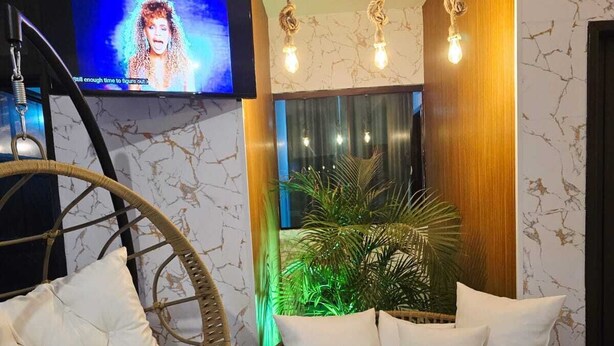Una habitación con un televisor montado en la pared, una silla de mimbre con cojines, una cama redonda de mimbre con almohadas y una planta en maceta.