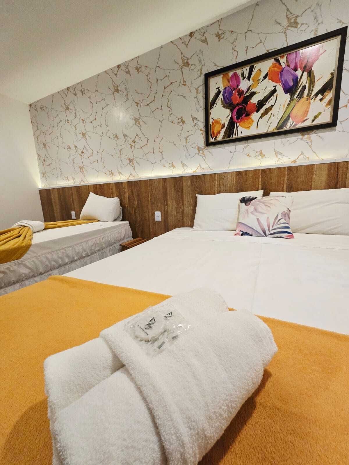 Habitación de hotel con una cama grande, una cama más pequeña, cabecera de madera, un cuadro de flores y una silla amarilla.