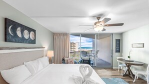 Habitació d'hotel amb llit, ventilador de sostre, balcó i vistes a la platja, i taula amb cadires.