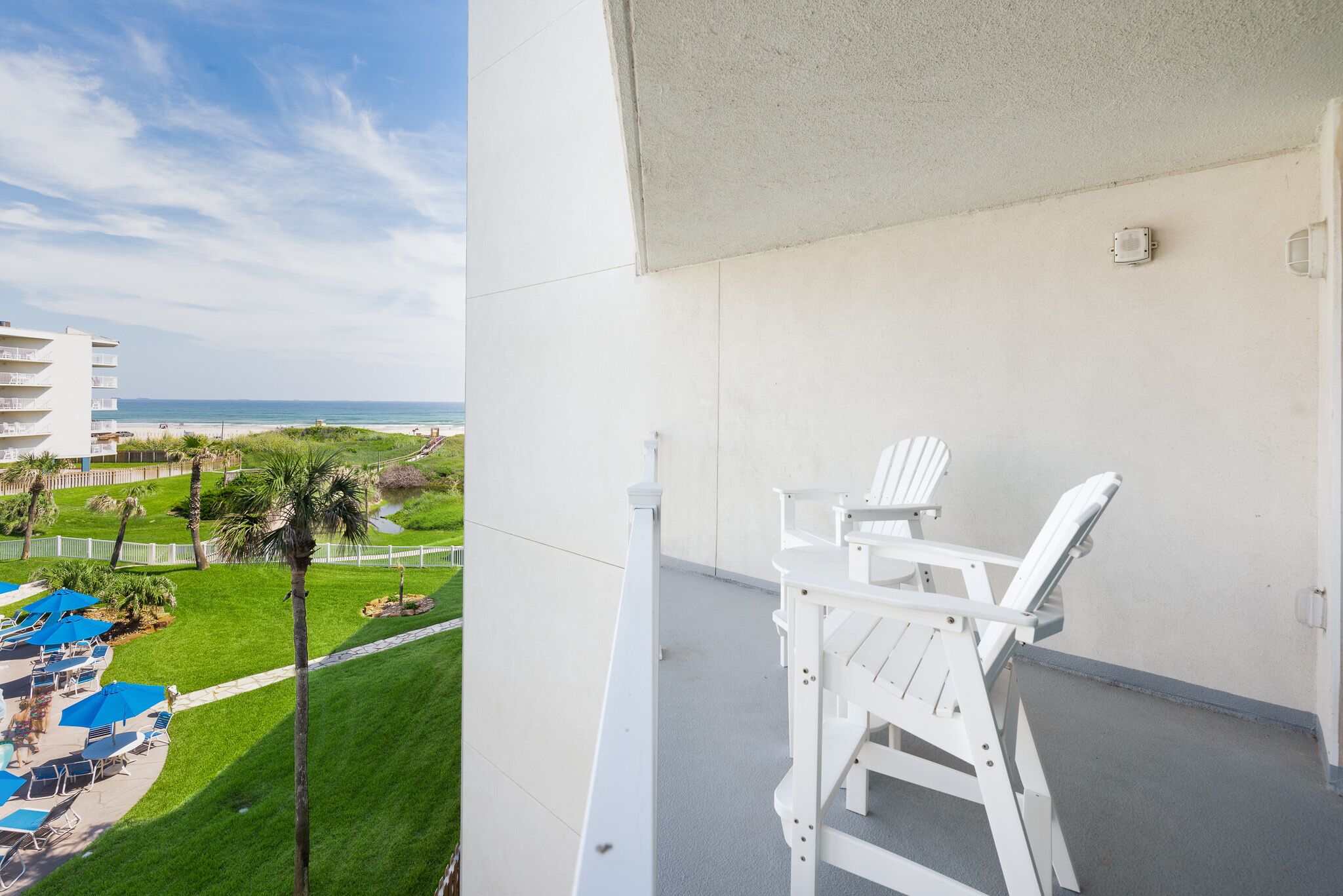 A balcony with white chairs and a view of a beach and resort area.