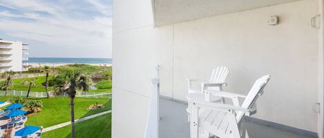 A balcony with white chairs and a view of a beach and resort area.
