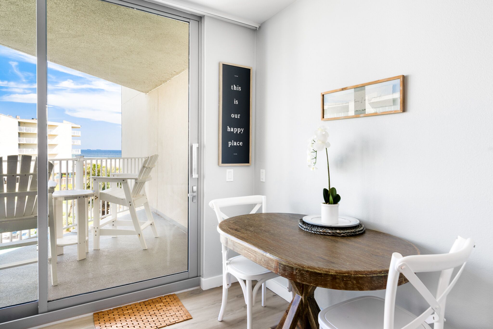 Un balcon avec vue sur l’océan, un coin repas avec une table et des chaises, et une citation inspirante encadrée au mur.