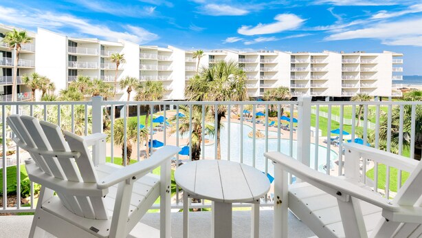 Un balcó amb cadires Adirondack blanques i una taula petita, amb vista a una zona de piscina amb palmeres i un edifici blanc al fons.
